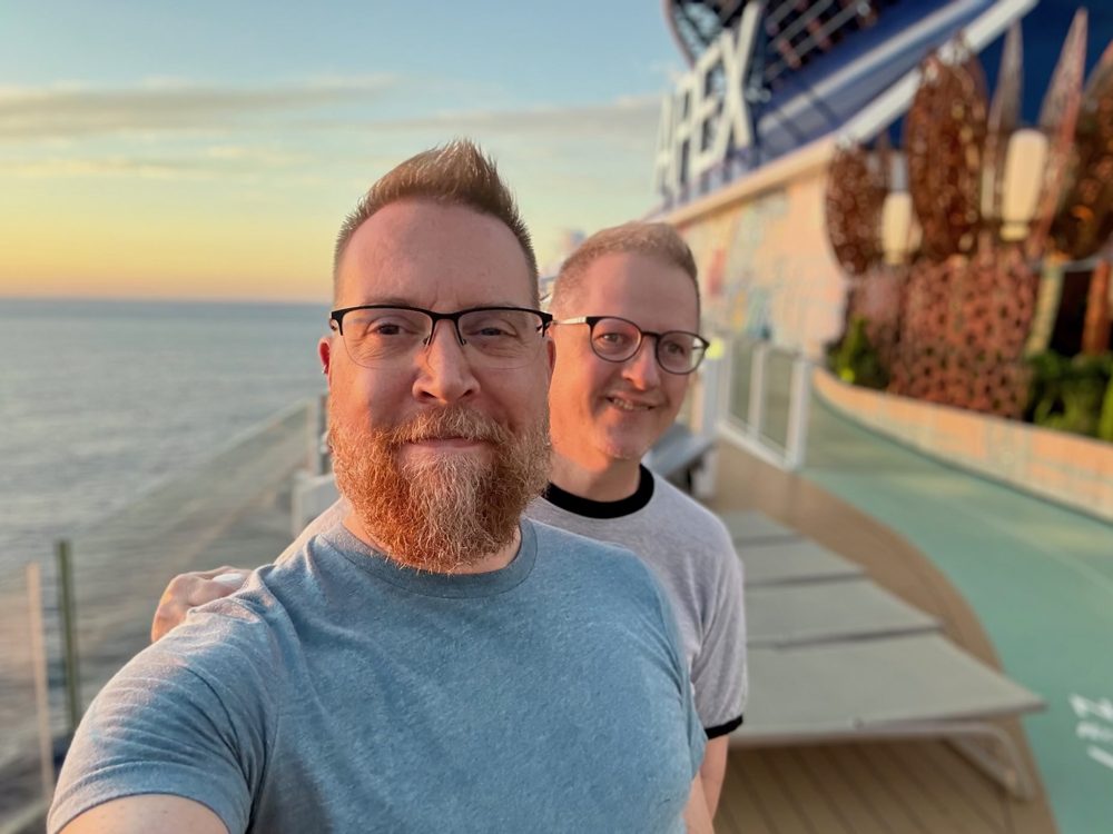 Andrew and his husband Joe smiling on the deck of a cruise ship at sunset